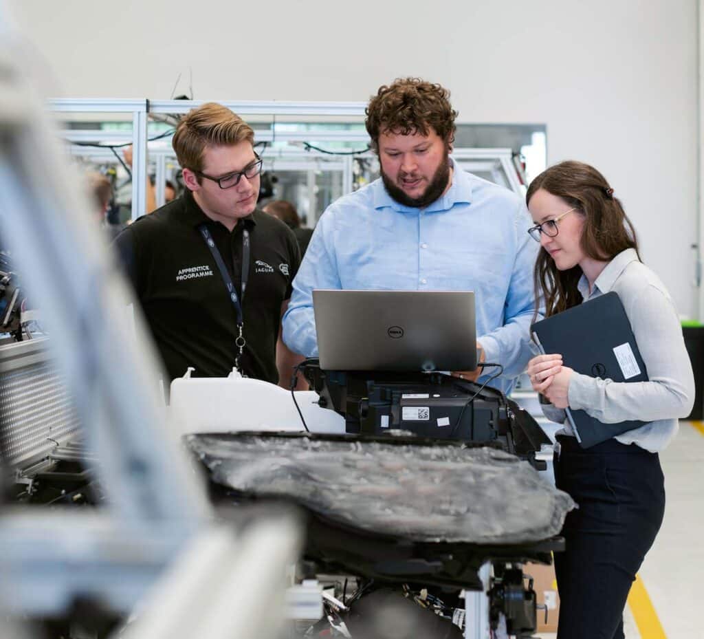 Drei Mitarbeiter in einem professionellen, industriellen Umfeld. Sie stehen an einem technischen Gerät, wobei eine Person an einem Laptop arbeitet.
